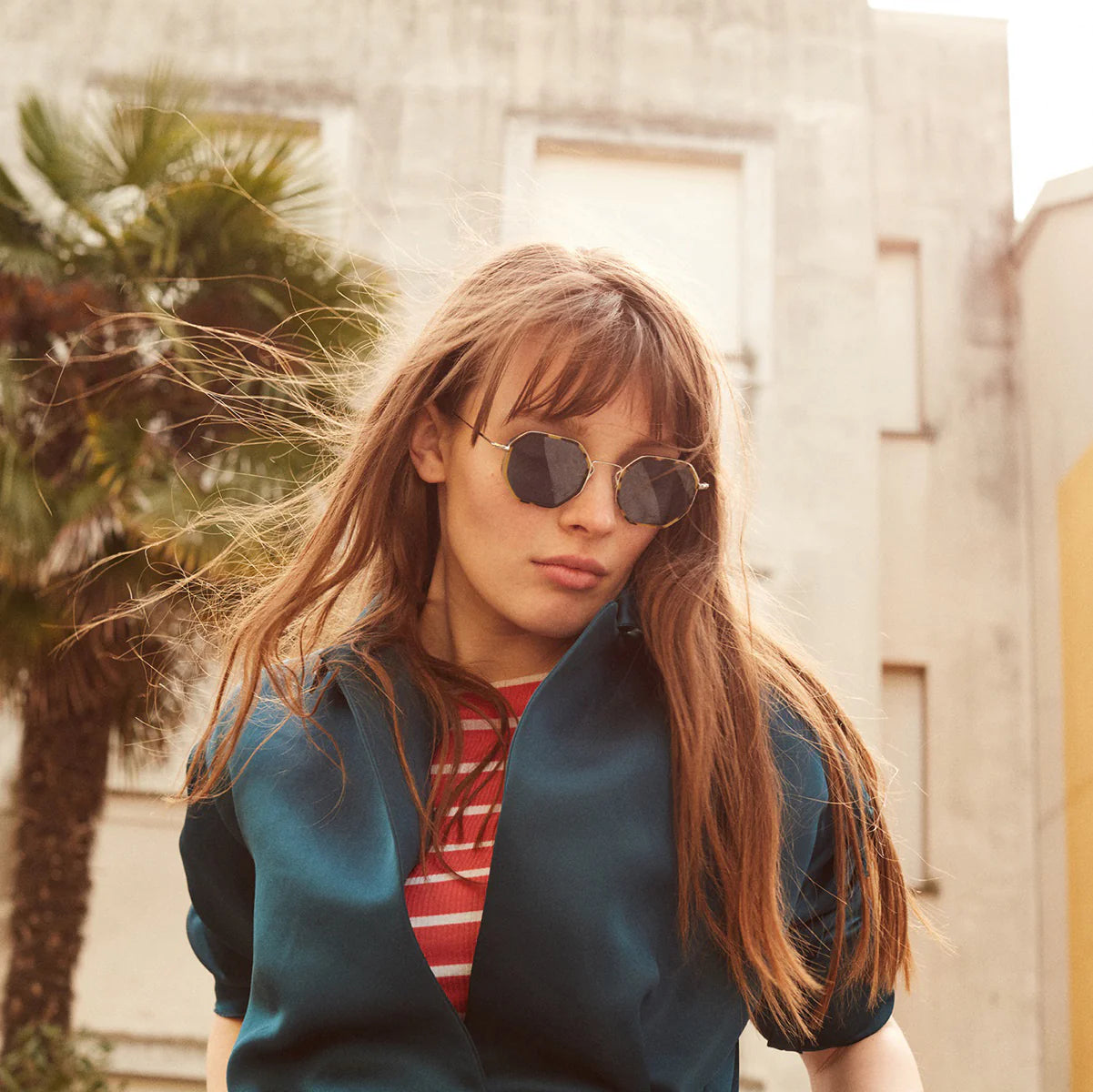 Woman wearing sunglasses and a blue jacket with a palm tree in the background