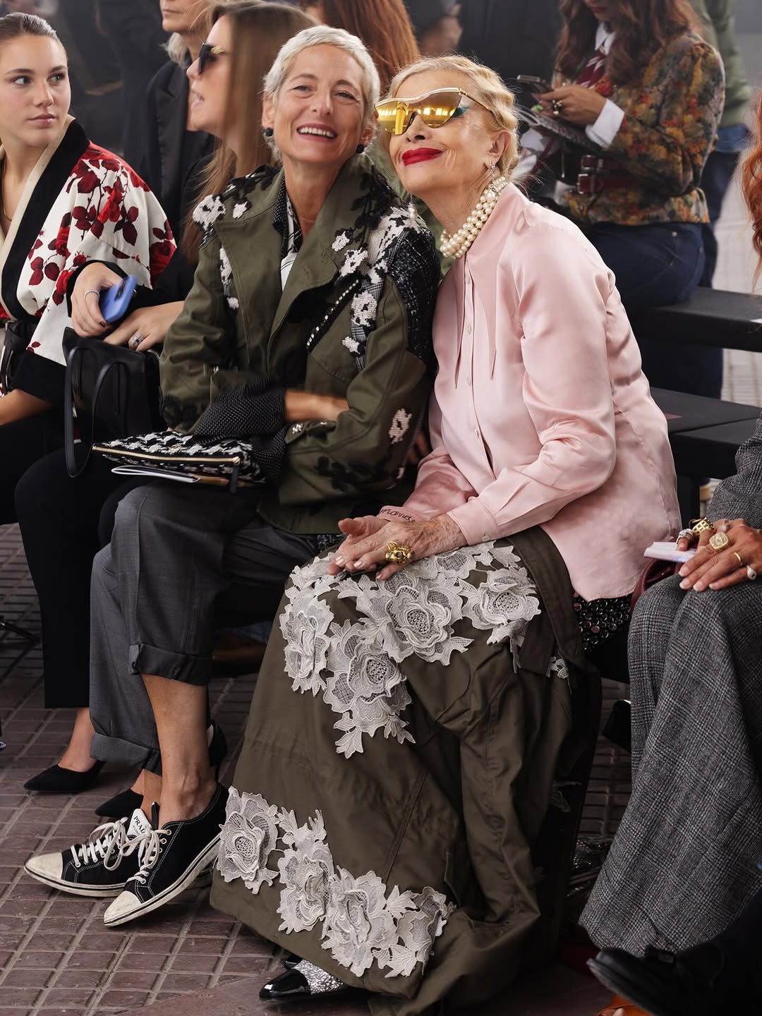 Two women sitting together at a fashion event, one wearing a floral dress and the other in a pink jacket.