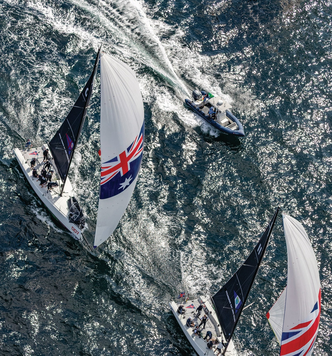 Two sailboats racing on a body of water with a small inflatable boat nearby.