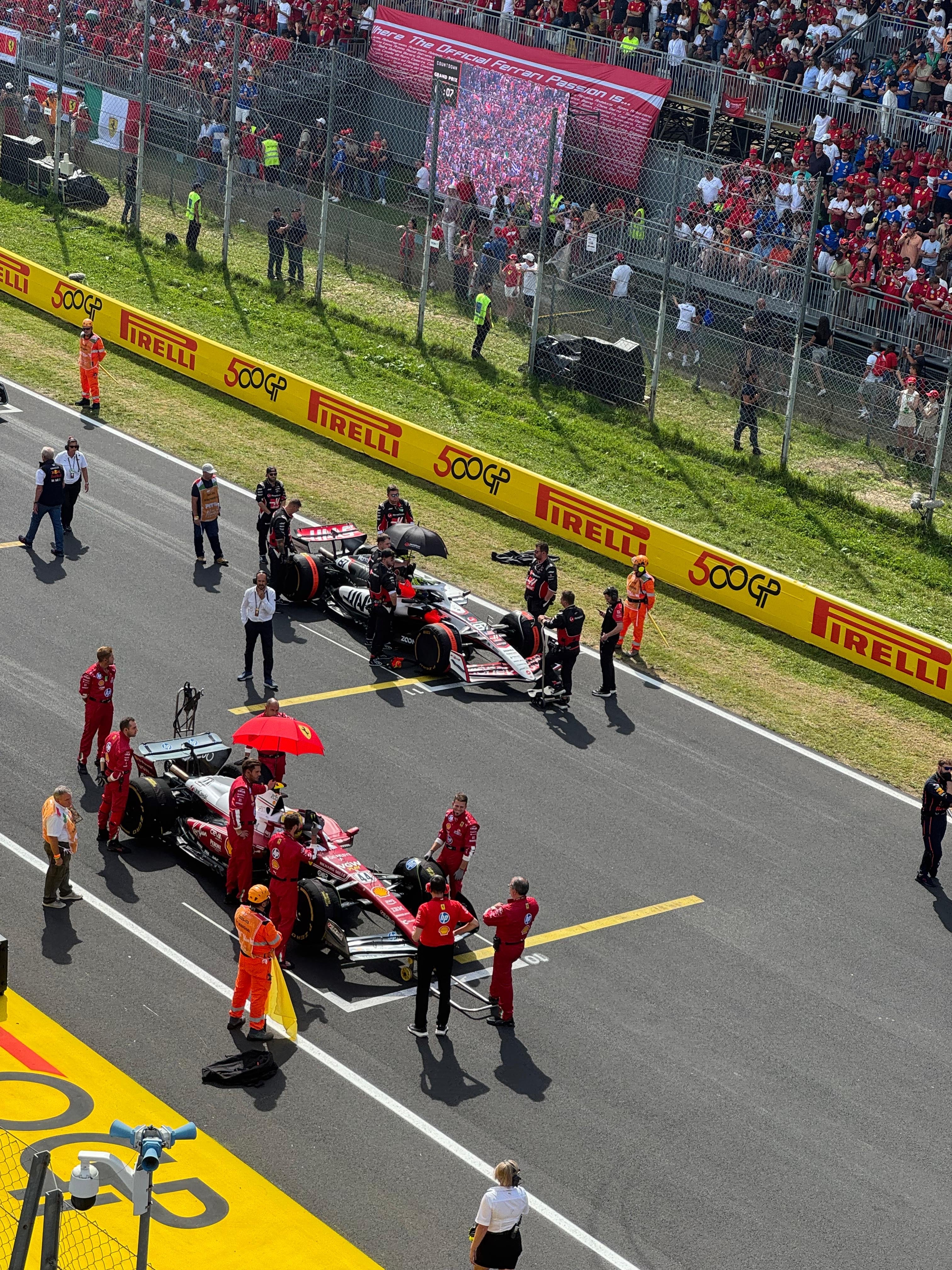 Ferrari Formula 1 pit stop on a racetrack with mechanics and cars, surrounded by spectators and Pirelli branding.