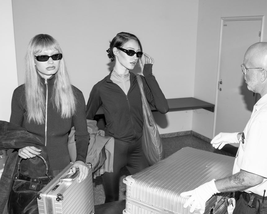 Two women in sunglasses with luggage, interacting with a man in a room.