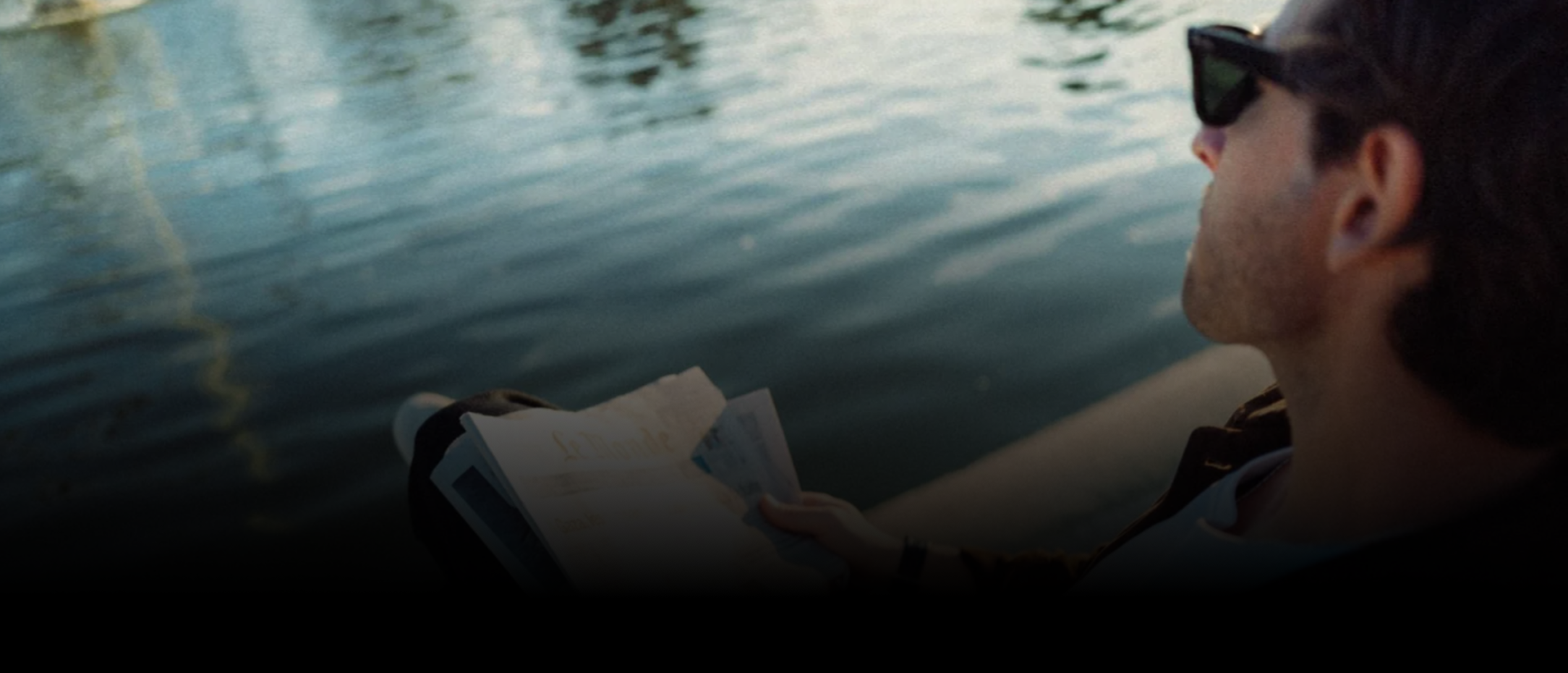 Person sitting by a body of water holding a book, wearing sunglasses.