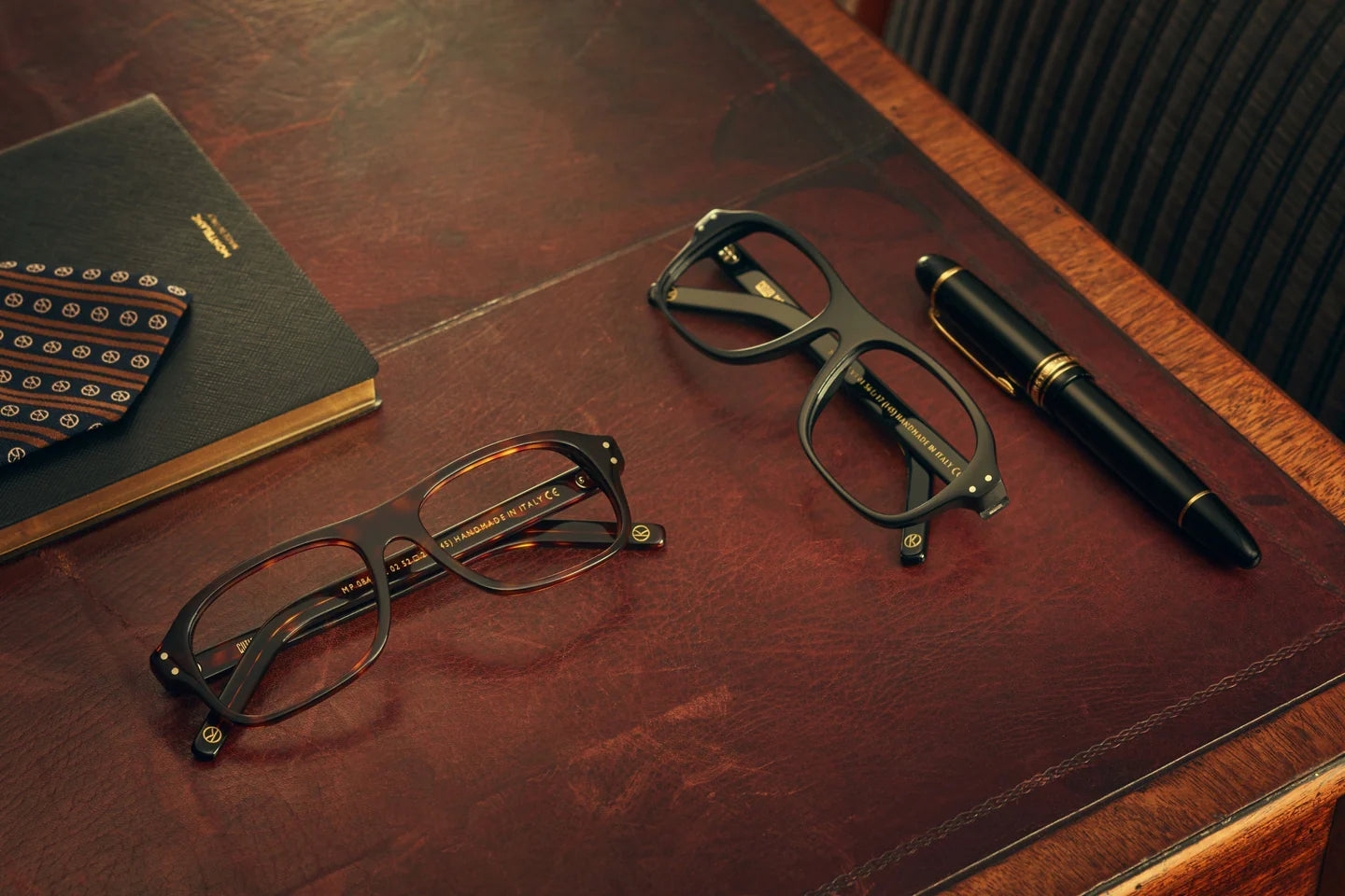 Two pairs of glasses and a pen on a wooden surface with a notebook.