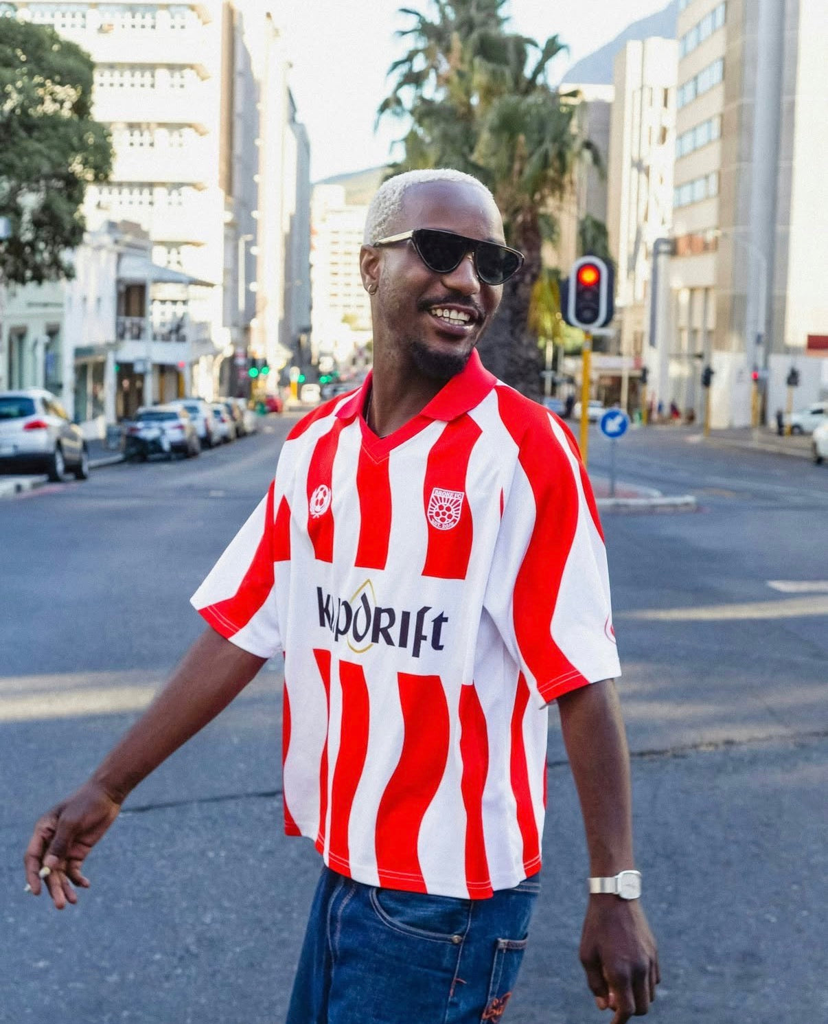 A man wearing a red and white vertically striped soccer jersey with 'Kodrift' printed across the chest, standing on a city street.