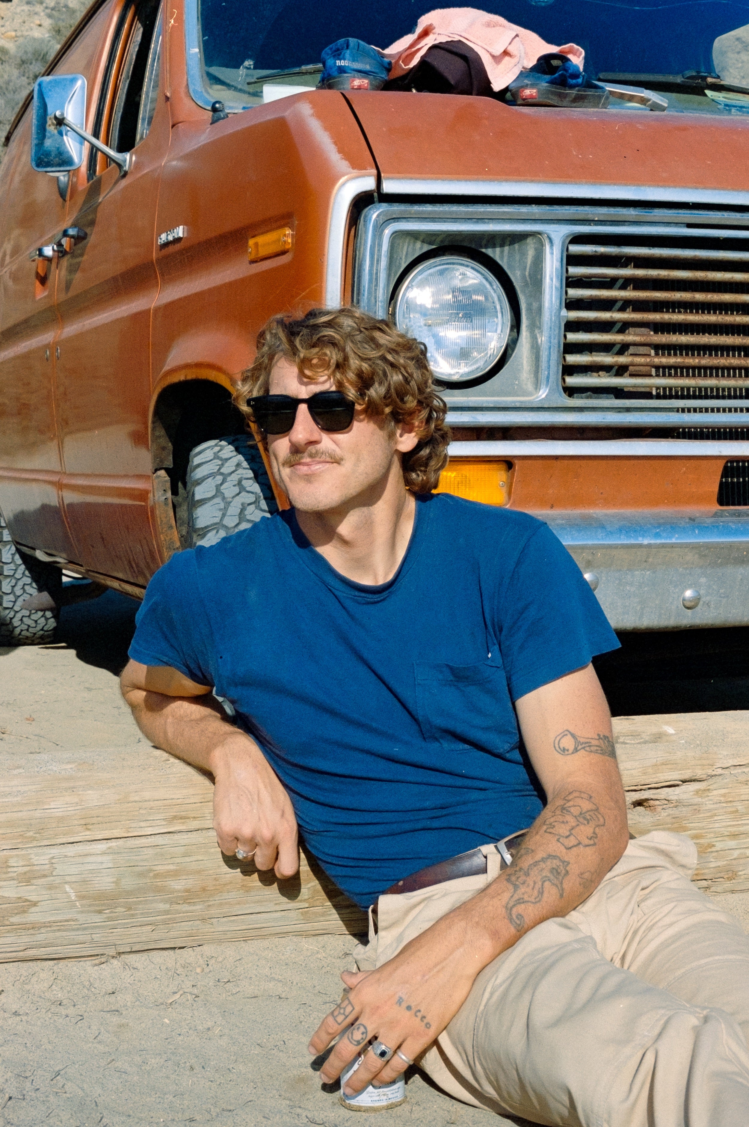 Man in sunglasses and blue shirt sitting in front of a rusted orange truck.