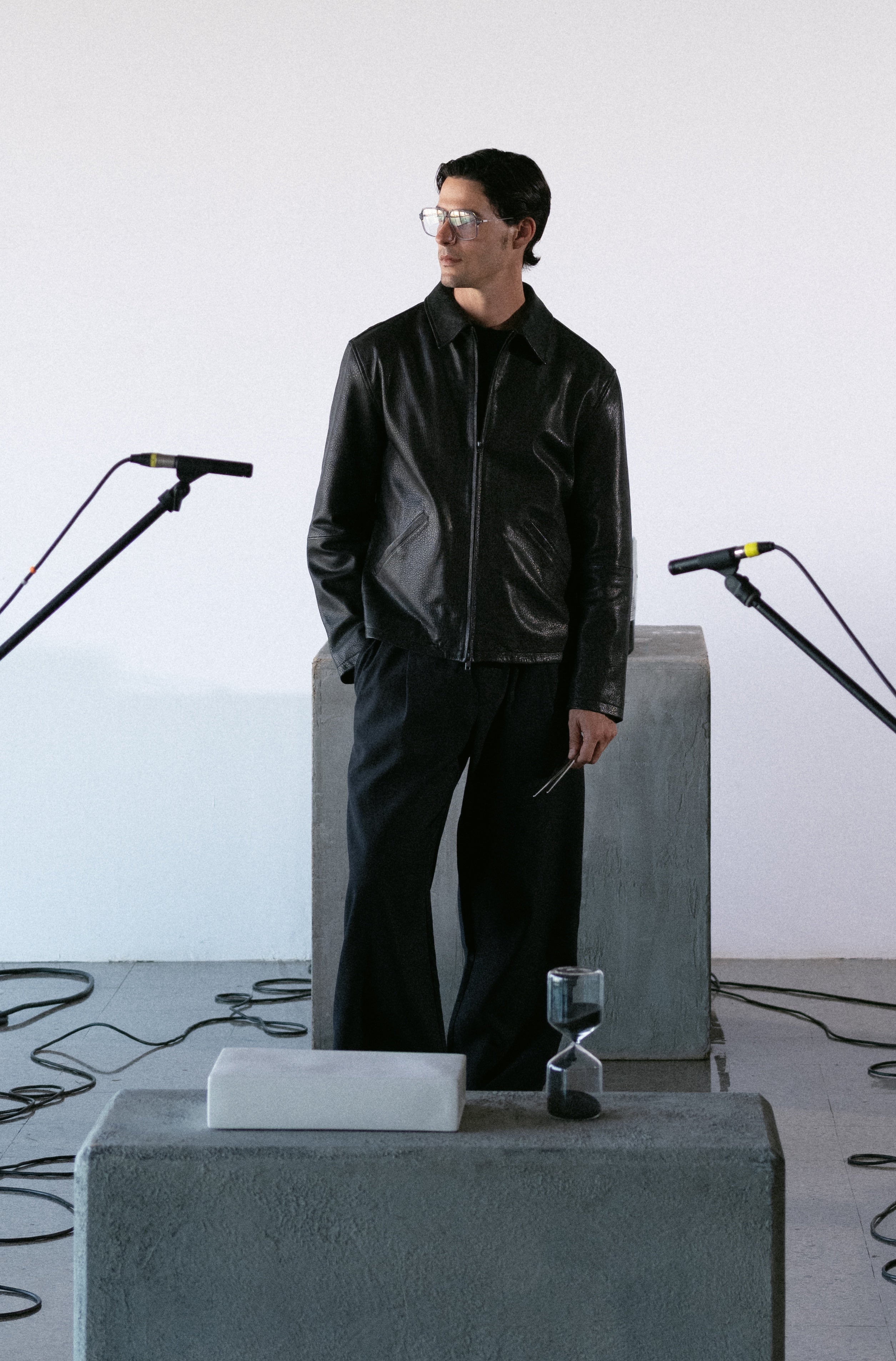 Person wearing a black leather jacket and pants in a minimalistic setting with concrete blocks and microphones.