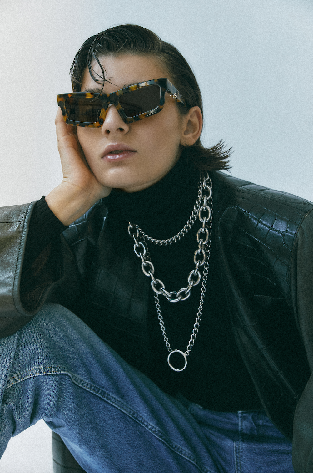 Person wearing sunglasses, black leather jacket, and blue jeans with a neutral background
