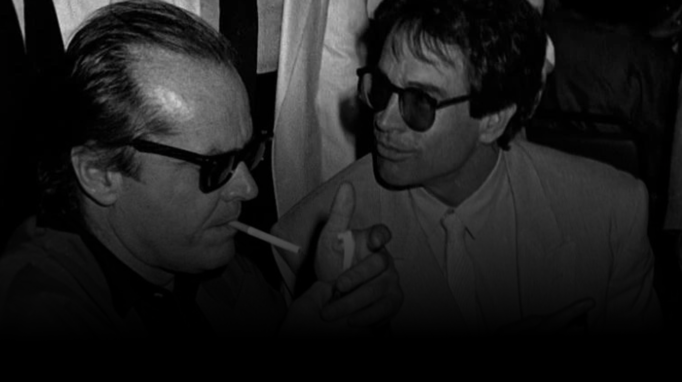 Two men in formal attire with sunglasses, one smoking a cigarette, in a black and white photo.