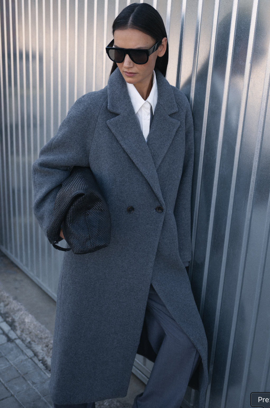 Person wearing a long gray coat and sunglasses against a metallic wall.