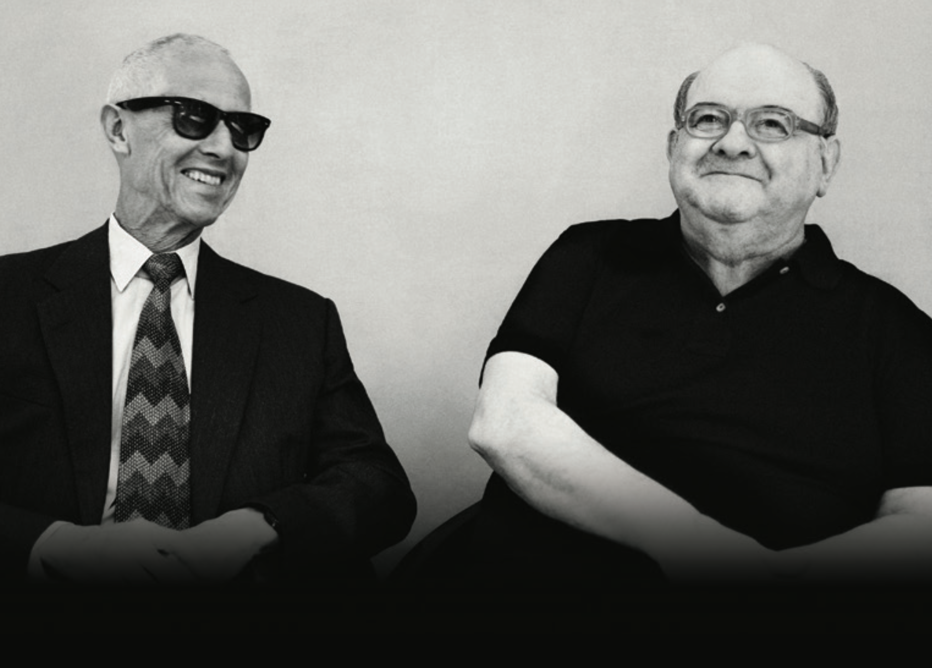 Two men sitting side by side against a plain background